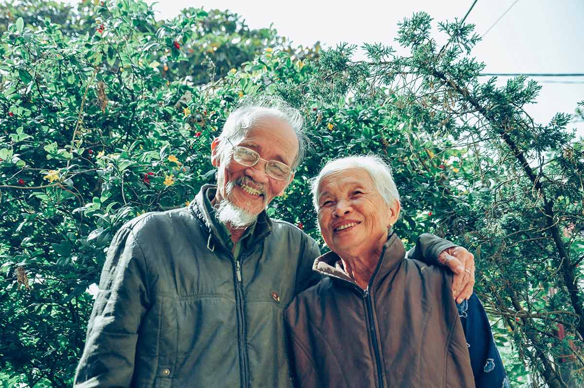 A happy old couple standing next to each other with trees in the background. 